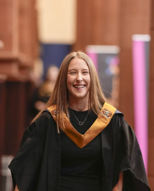 A cheerful female graduate walking down the aisle in her black gown and gold stole at the ceremony. A cheerful female graduate walking down the aisle in her black gown and gold stole at the ceremony.